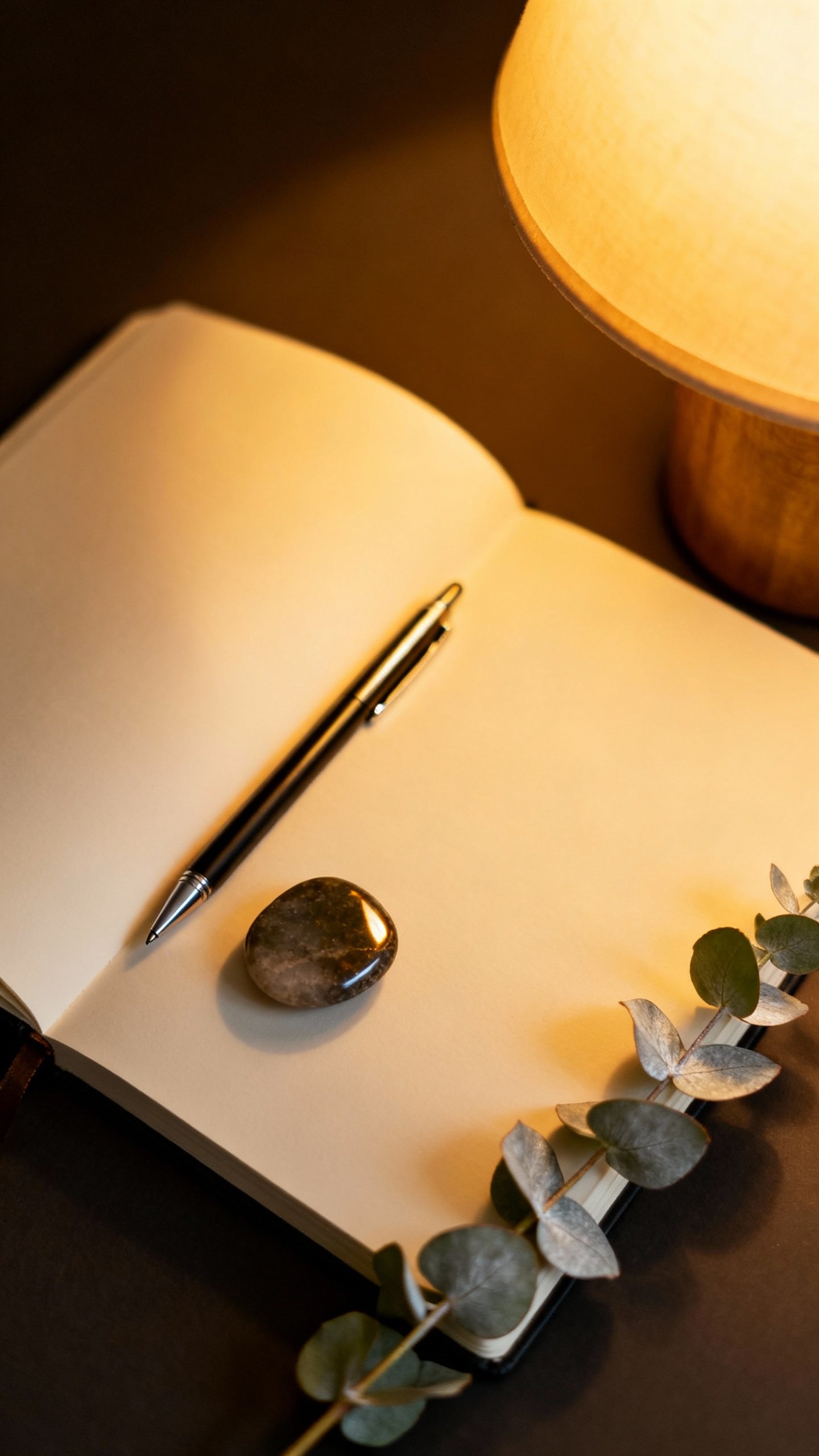Journaling corner detail: open notebook, fine-tip pen, polished stone token, eucalyptus sprig, dim l