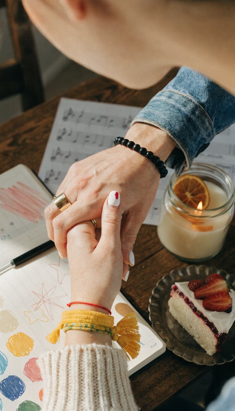 Quirk-forward couple moment, candid and warm: a close-up of two hands entwined over a planner filled with doodles and color swatches, sunlight catching a ring band in the foreground. The surrounding scene includes a softly blurred playlist paper, a tiny cake slice on a plate, and a citrus-scented candle. The person in the scene is present but not the focus—back of a head or a shoulder visible, slightly out of focus.
