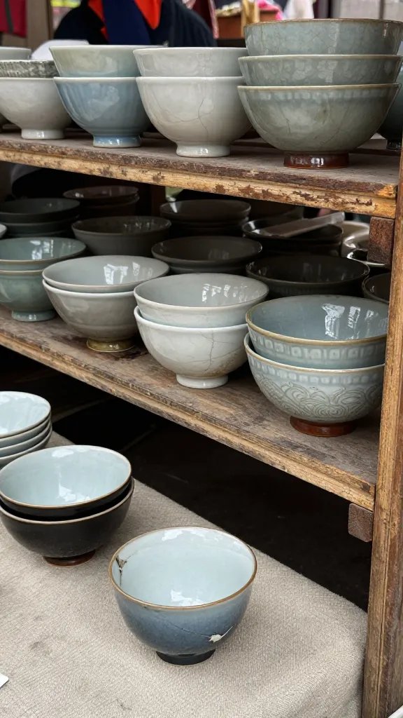 A candid market scene framed to emphasize the interior story: a seller’s worn shelf of bowls and cups with varying glaze behaviors (craze lines, glaze sag, and even wear). The foreground features a chipped teacup resting on a beige cloth, inviting the viewer to distinguish quality; a distant shopper is blurred to keep the pieces as the focal point.