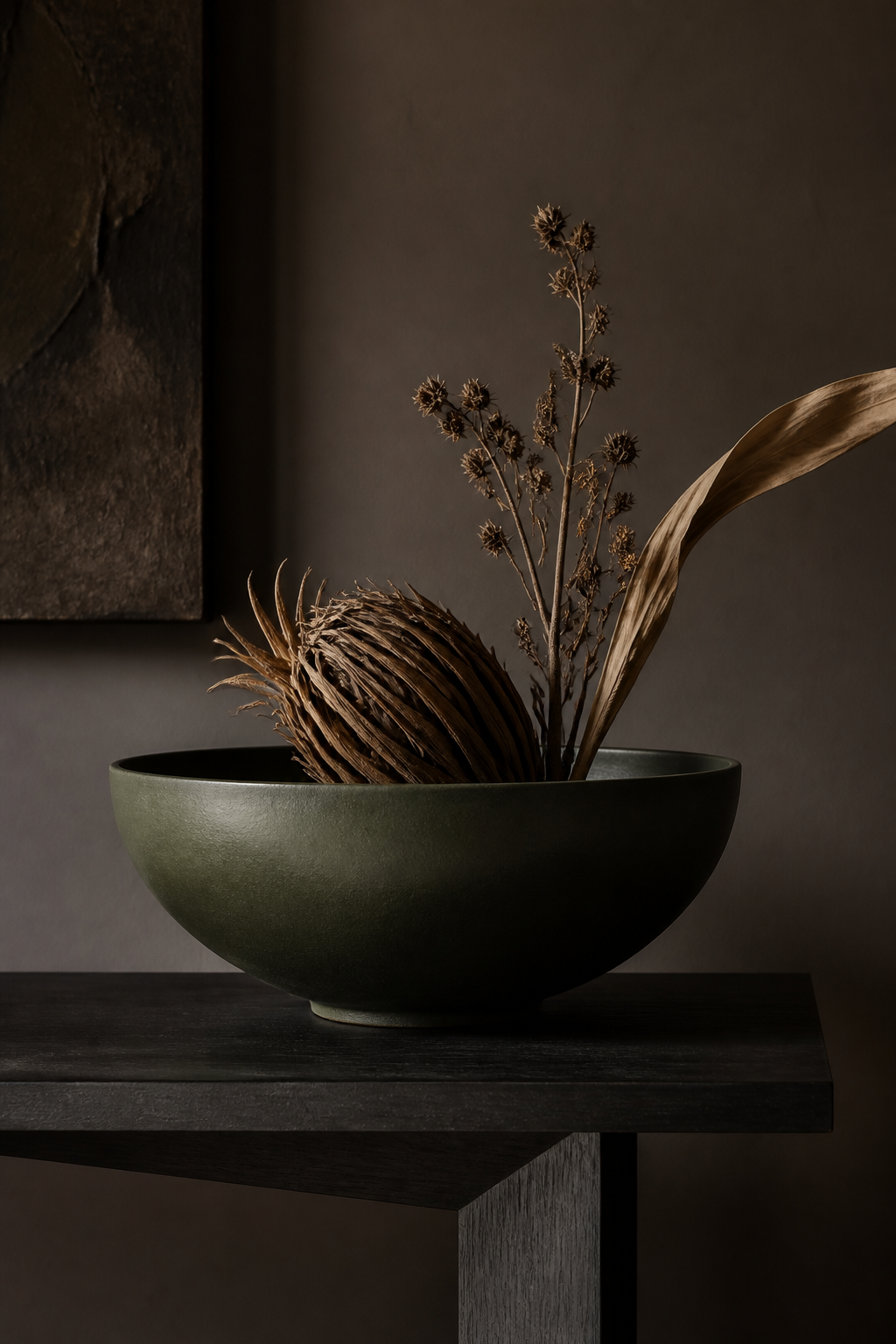 A close-up vignette on a minimalist console table made of dark wood with a matte black finish. It features a single, oversized bowl in a deep olive tone, holding a few dried, architectural botanical elements. The background wall is painted in a sophisticated smoky taupe, accentuating the depth and luxurious choice of objects. No people, no text.