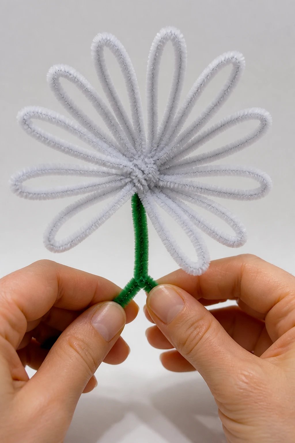 A photorealistic 16:9 studio shot of a single pipe cleaner daisy being assembled: multiple white pipe cleaners forming slender petals around a central core, with a visible twist technique and a faint shadow to emphasize texture, on a clean neutral backdrop.